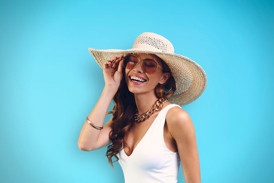 A stylish woman wearing a chunky gold necklace, paired with a white outfit and a wide-brimmed straw hat, exuding summer elegance against a bright blue background - Mia Ishaaq