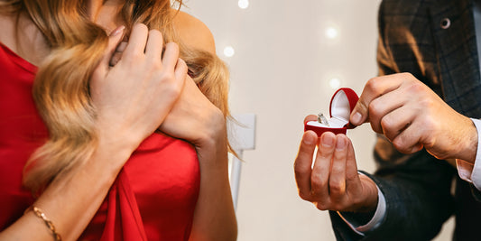 A romantic scene of a surprise marriage proposal, featuring a man presenting an engagement ring in a red velvet box to a woman in a red dress - Mia Ishaaq