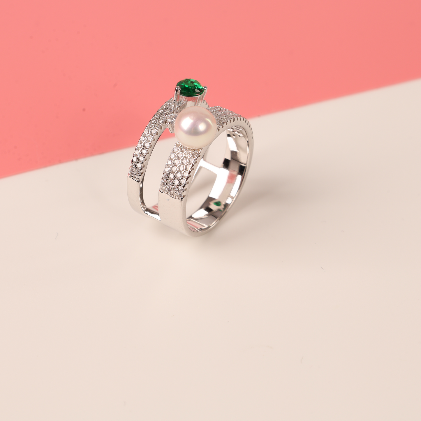 The Green Cubic Zirconia Pearl Ring by Mia Ishaaq, crafted in sterling silver with a lustrous pearl and accented with emerald-like stones and diamonds, rests elegantly on a pink and white background.
