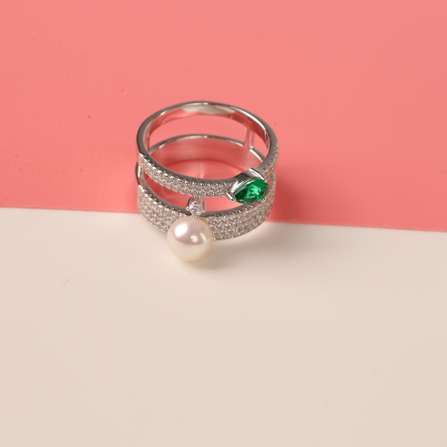 The Green Cubic Zirconia Pearl Ring by Mia Ishaaq, crafted in sterling silver and accented with a pearl and green cubic zirconia, is stylishly set against a soft pink and white backdrop.