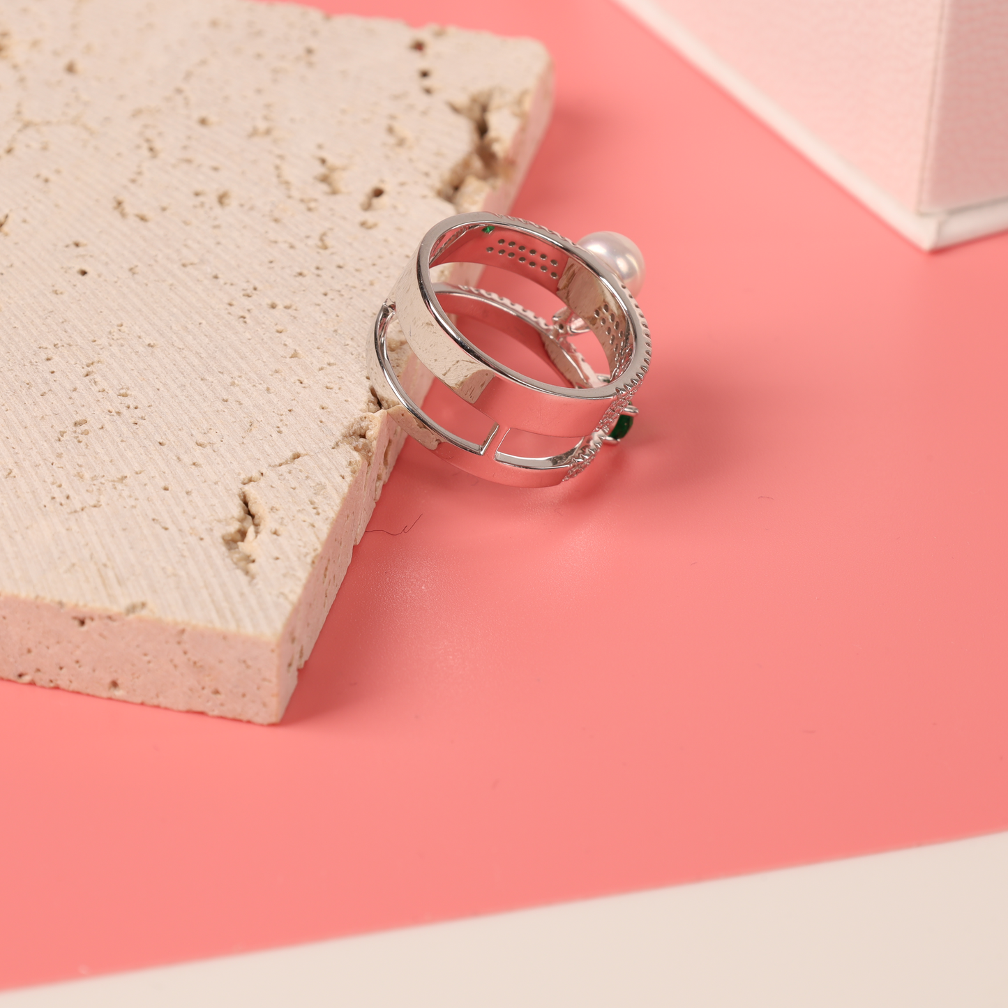 The Mia Ishaaq Green Cubic Zirconia Pearl Ring gracefully sits on a beige stone slab against a pink background, exuding timeless beauty.
