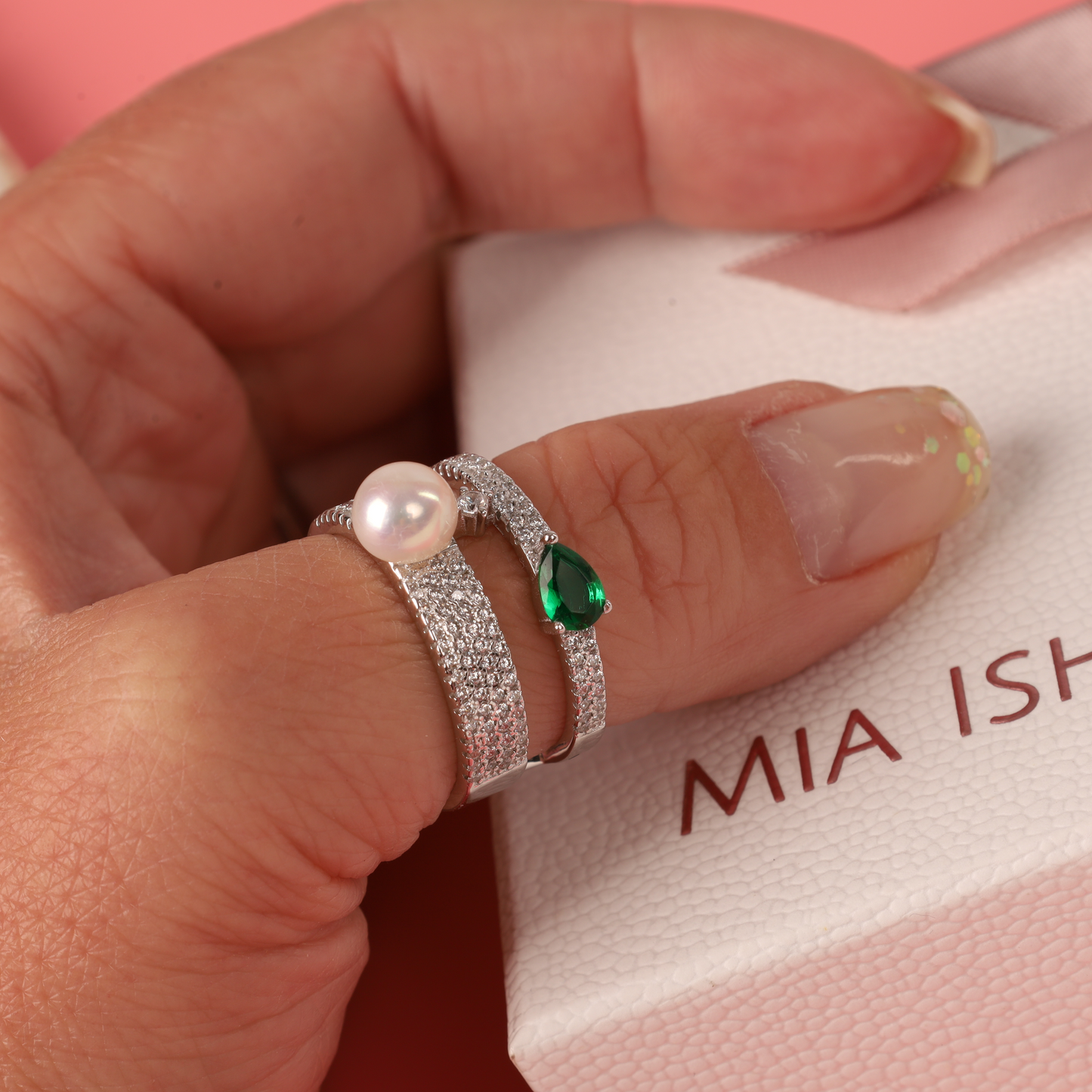 A hand adorned with Mia Ishaaq's Green Cubic Zirconia Pearl Ring in sterling silver rests gracefully on a textured box, set against a pink backdrop.