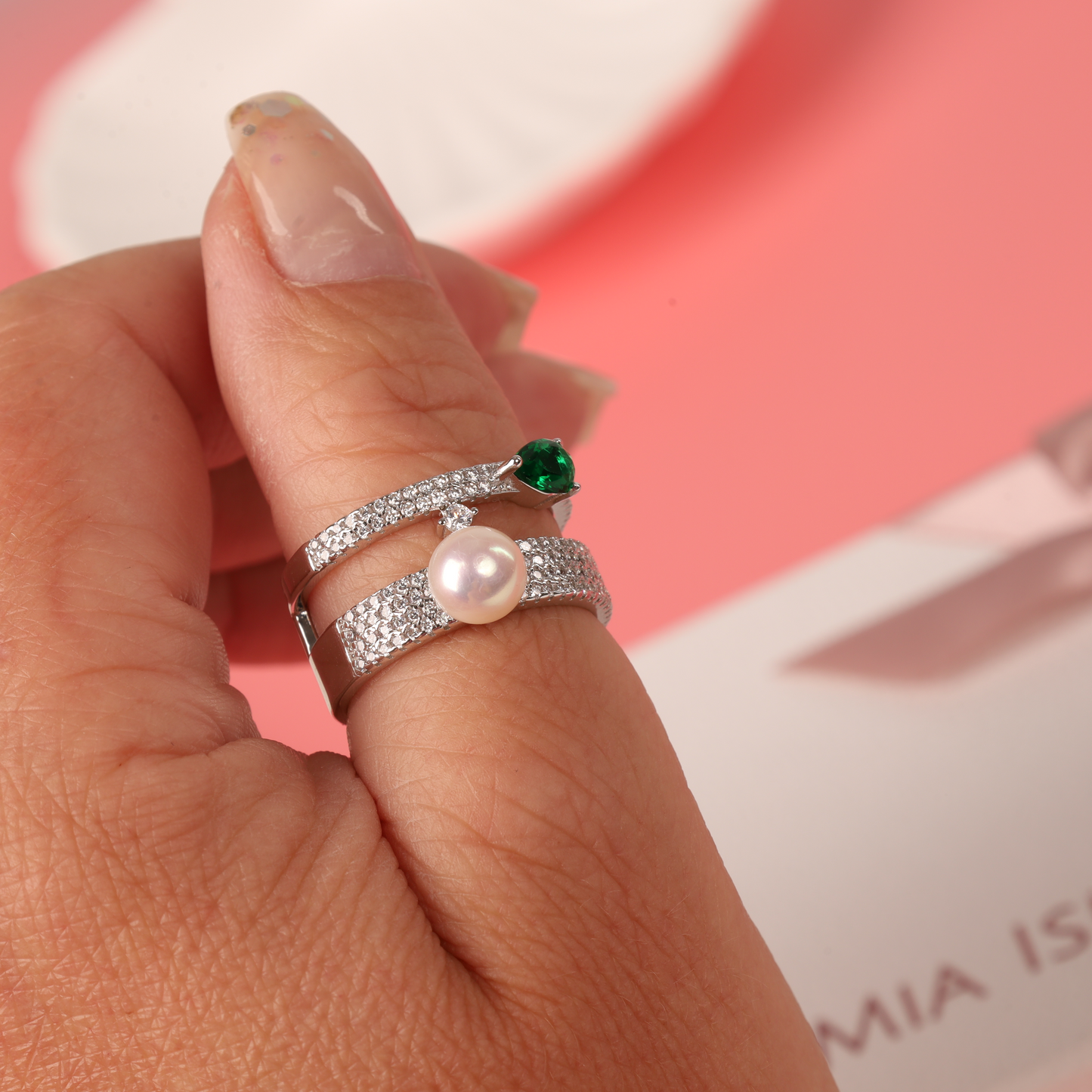 A hand adorned with Mia Ishaaq's Green Cubic Zirconia Pearl Ring, featuring a glowing pearl and a pop of green, shines against a pink background.