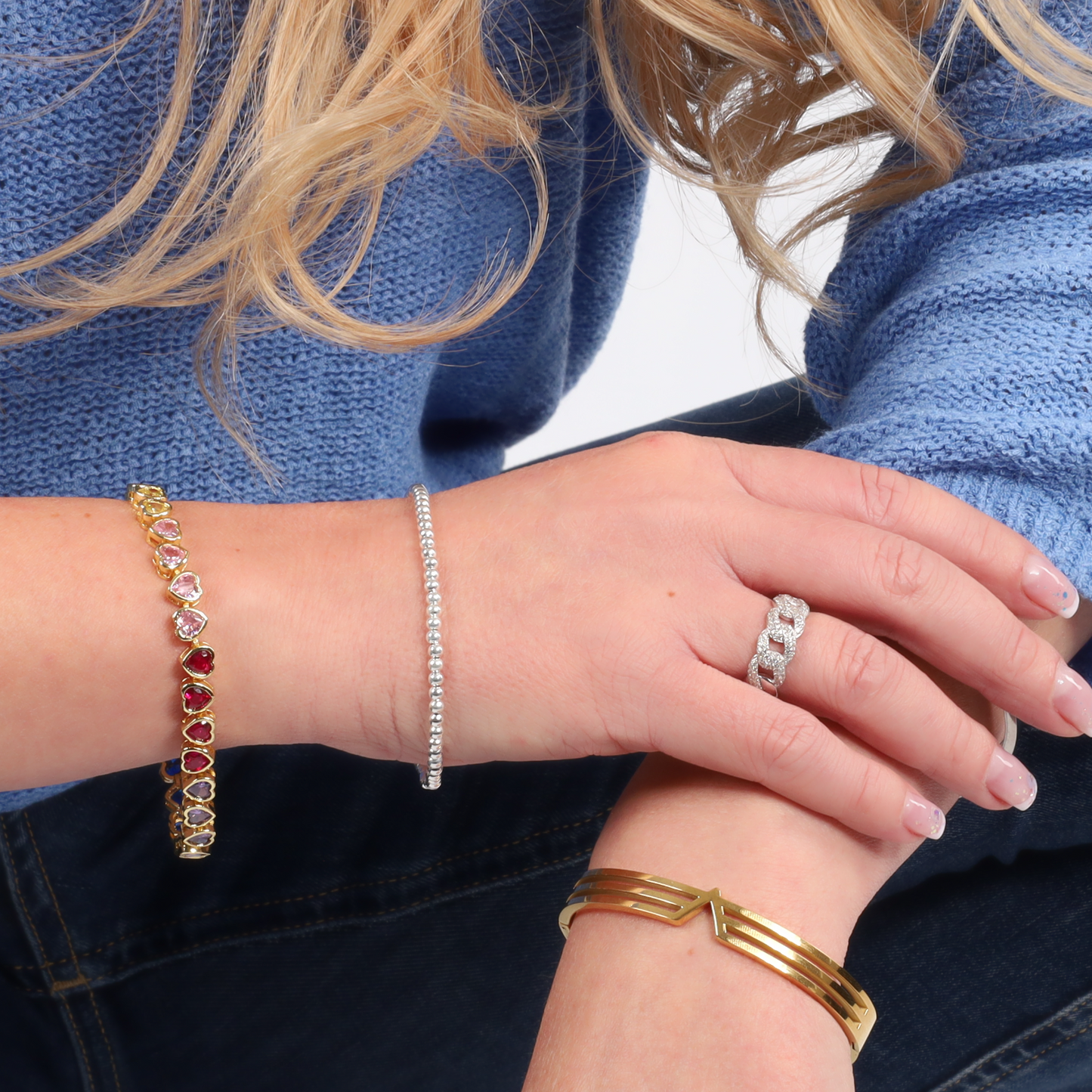 A close-up shot features a person wearing three bracelets and a Mia Ishaaq Cuban Link Chain Ring embellished with cubic zirconia stones, hands crossed. They are dressed in blue with long, wavy hair.