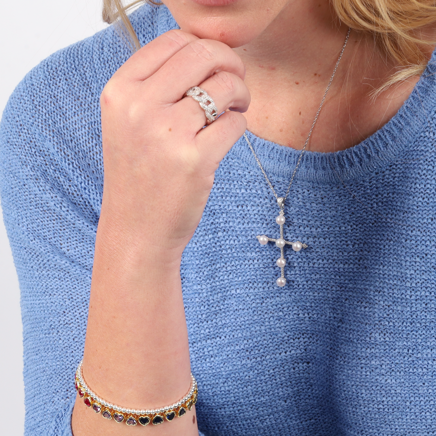 A person in a blue sweater, adorned with a cross necklace, silver rings, and a colorful bracelet, complements their hand with the Cuban Link Chain Ring featuring cubic zirconia stones by Mia Ishaaq.