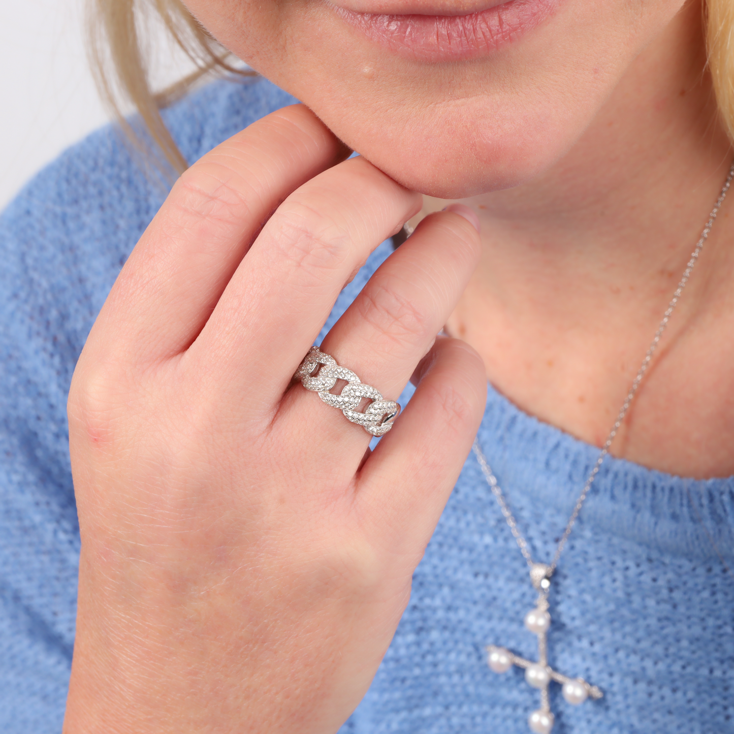 A woman in a blue sweater wearing a Cuban Link Chain Ring by Mia Ishaaq and a pearl cross necklace, touching her face.