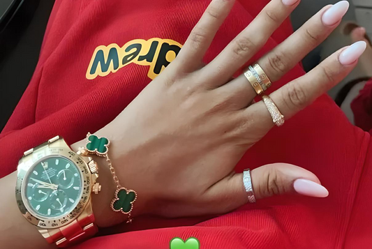 A close-up of a hand adorned with elegant jewelry, including gold rings, a green clover bracelet, and a gold watch with a green face - Mia Ishaaq