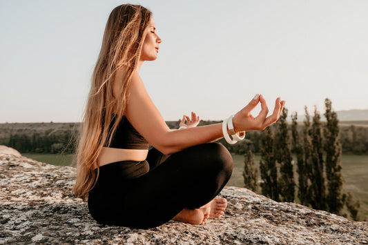 A woman practicing yoga outdoors on a rocky surface, wearing a black sports outfit and white bangles, with a scenic natural background - Mia Ishaaq