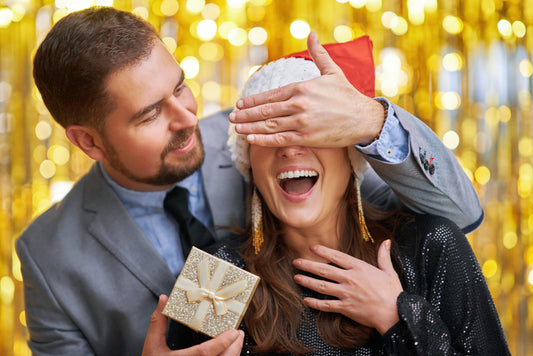 Man holding gold coloured gift wrap box while covering a woman's eyes with his hand - Mia Ishaaq