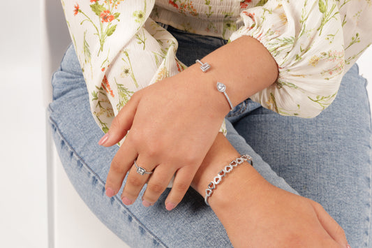 Model sitting down on a white chairs with her arms resting on her leg wearing a silver heart bangle bracelet on one wrist and open bangle bracelet with cubic zirconia on the other wrist - Mia Ishaaq