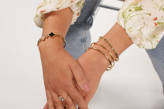 Model sitting on a white chair with her hands slightly crossed over and wearing stack of gold bangle bracelets on one hand and four leaf bangle bracelet on the other hand - Mia Ishaaq