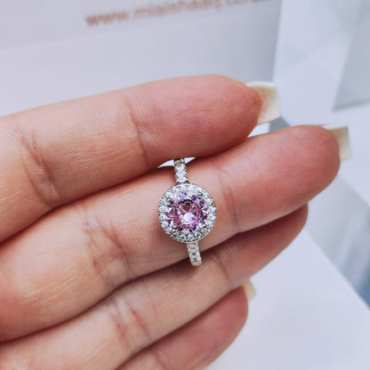 Model holding the ring delicately between their fingers, allowing a clear view of the pink cubic zirconia and surrounding halo stones - Mia Ishaaq