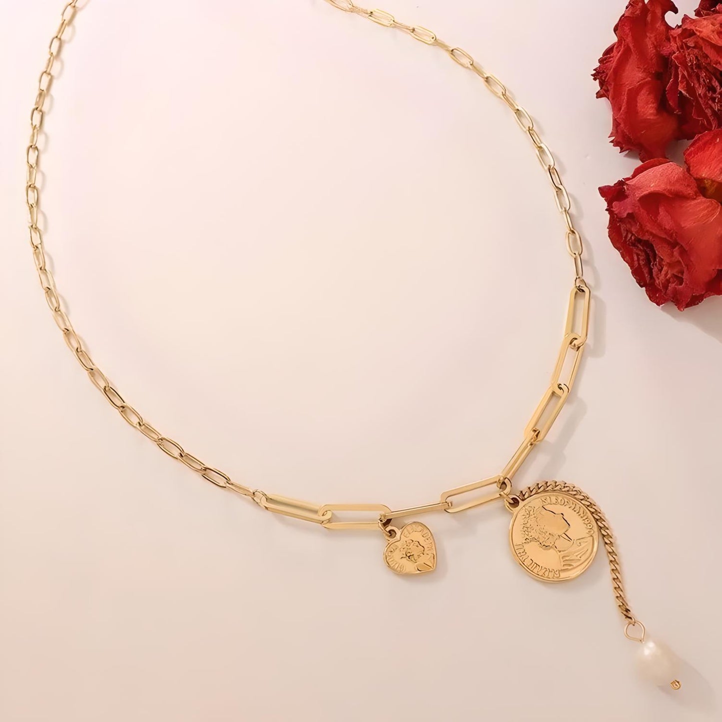 The Pearl & Coin Charm Link Necklace by Mia Ishaaq, featuring 18K gold plating with pearl and coin charm accents, is beautifully showcased alongside red dried flowers on a white background.