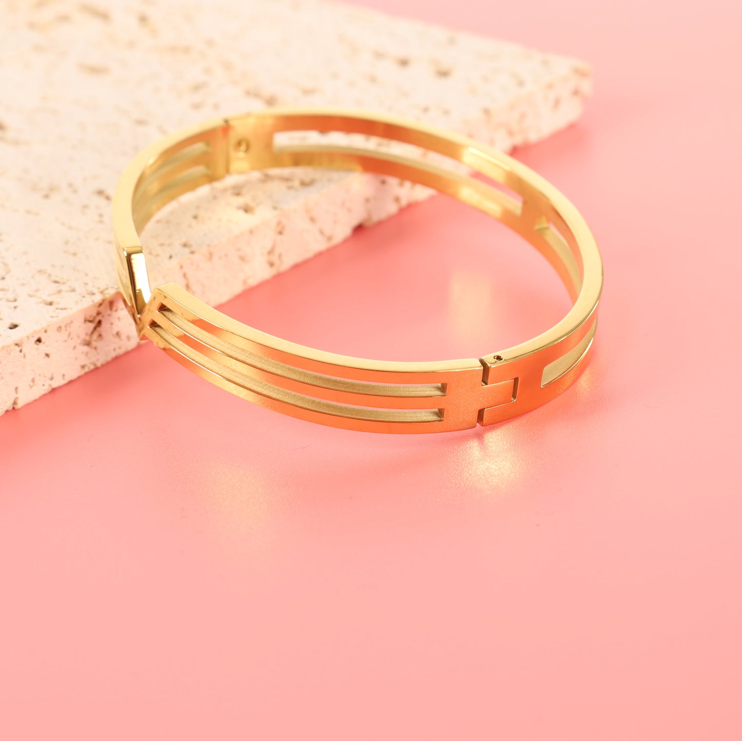 The Mia Ishaaq Gold V Line Harmony Bangle Bracelet, featuring 18k gold plating with intricate cut-out geometric designs, rests on a light pink surface.