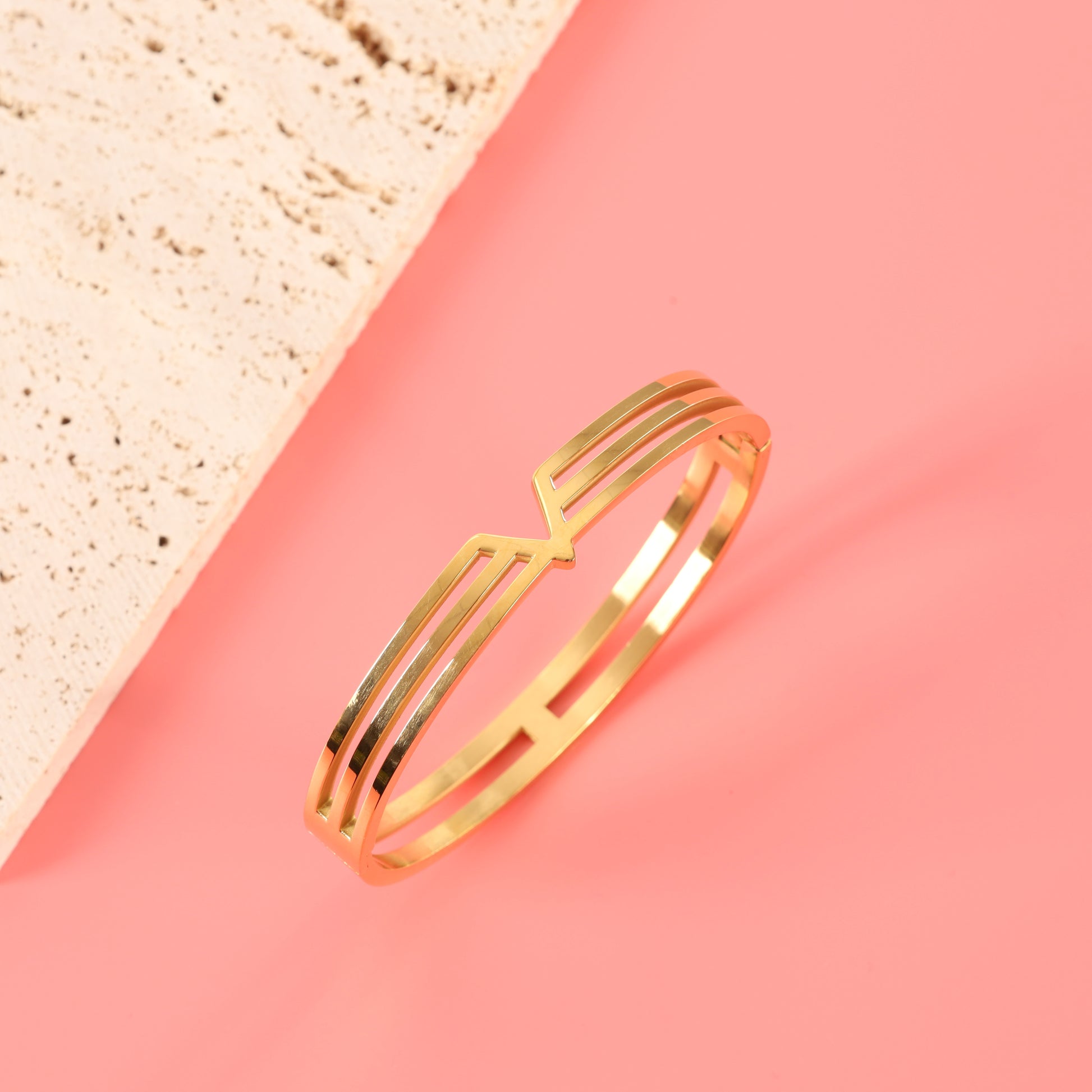 Mia Ishaaq Gold V Line Harmony Bangle Bracelet in 18k PVD Gold, minimalist V-shaped design, displayed against a pink background.