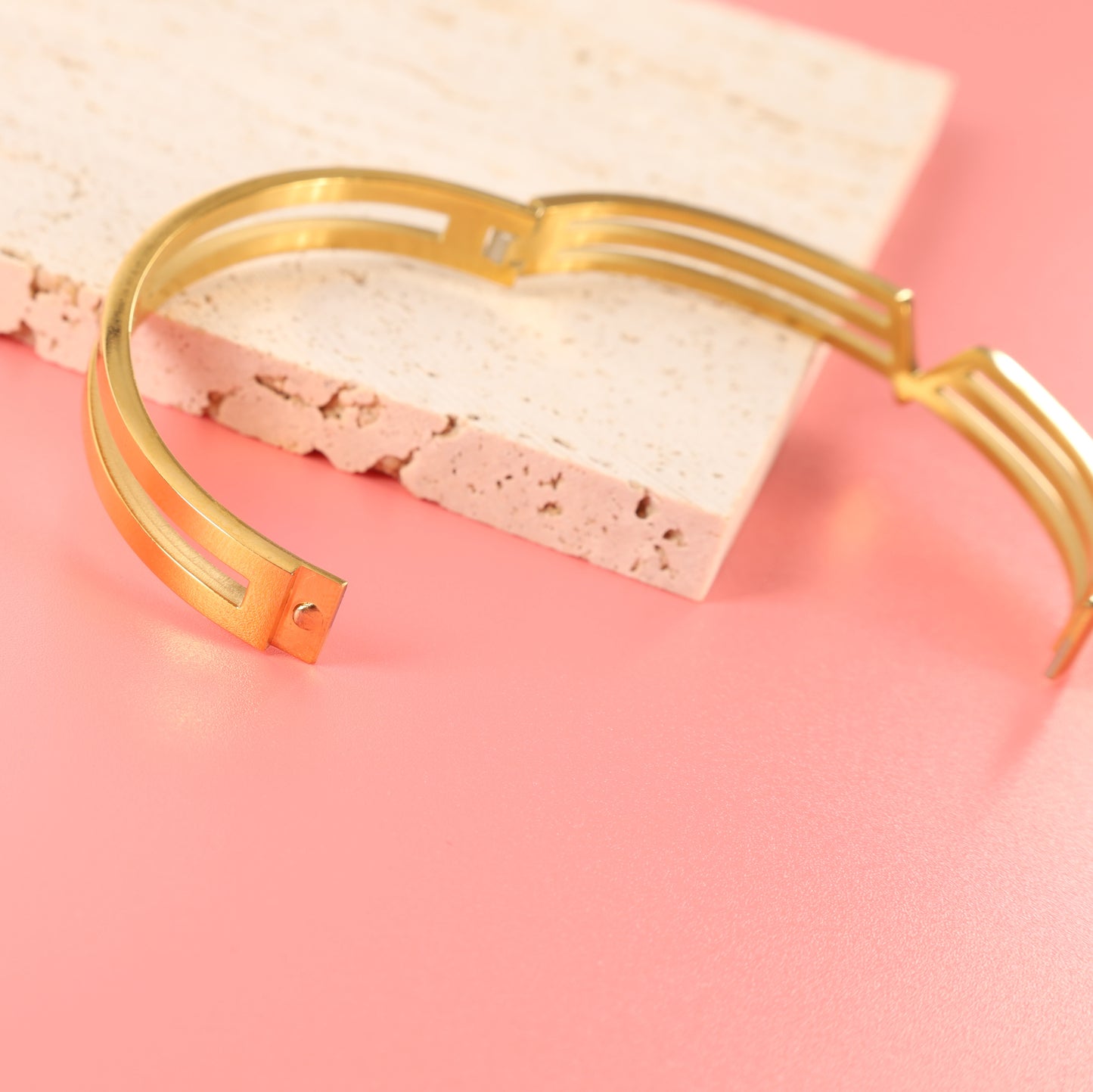 Mia Ishaaq Gold V Line Harmony Bangle Bracelet, resting on a textured white stone slab against a soft pink background.