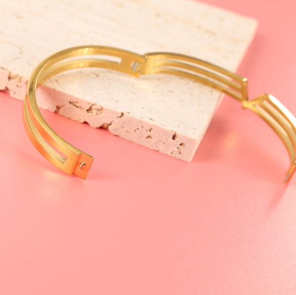 Mia Ishaaq Gold V Line Harmony Bangle Bracelet, resting on a textured white stone slab against a soft pink background.