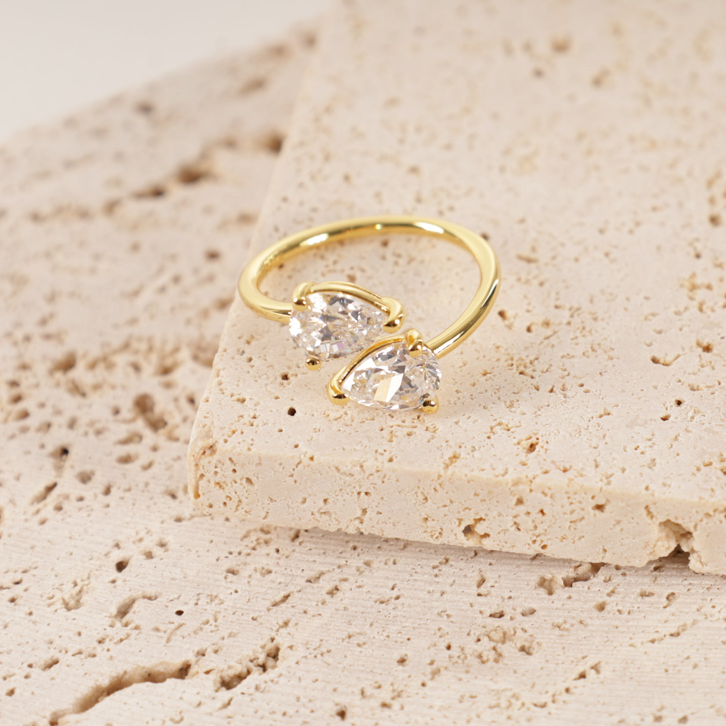 A Gold Twist Teardrop Ring by Mia Ishaaq, plated in 14k gold and adorned with two teardrop cubic zirconia stones, rests elegantly on a textured stone surface.