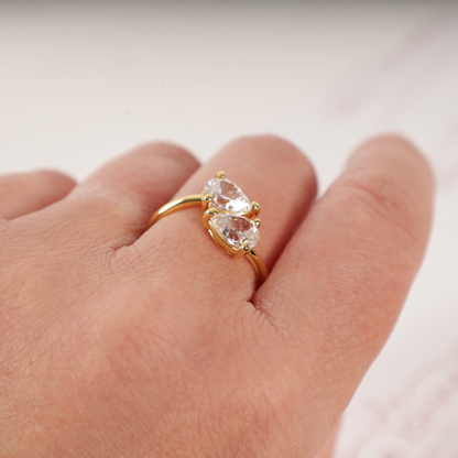 A hand adorned with the Mia Ishaaq Gold Twist Teardrop Ring, featuring two oval-shaped clear stones and a 14k gold-plated finish, is set against a light background.