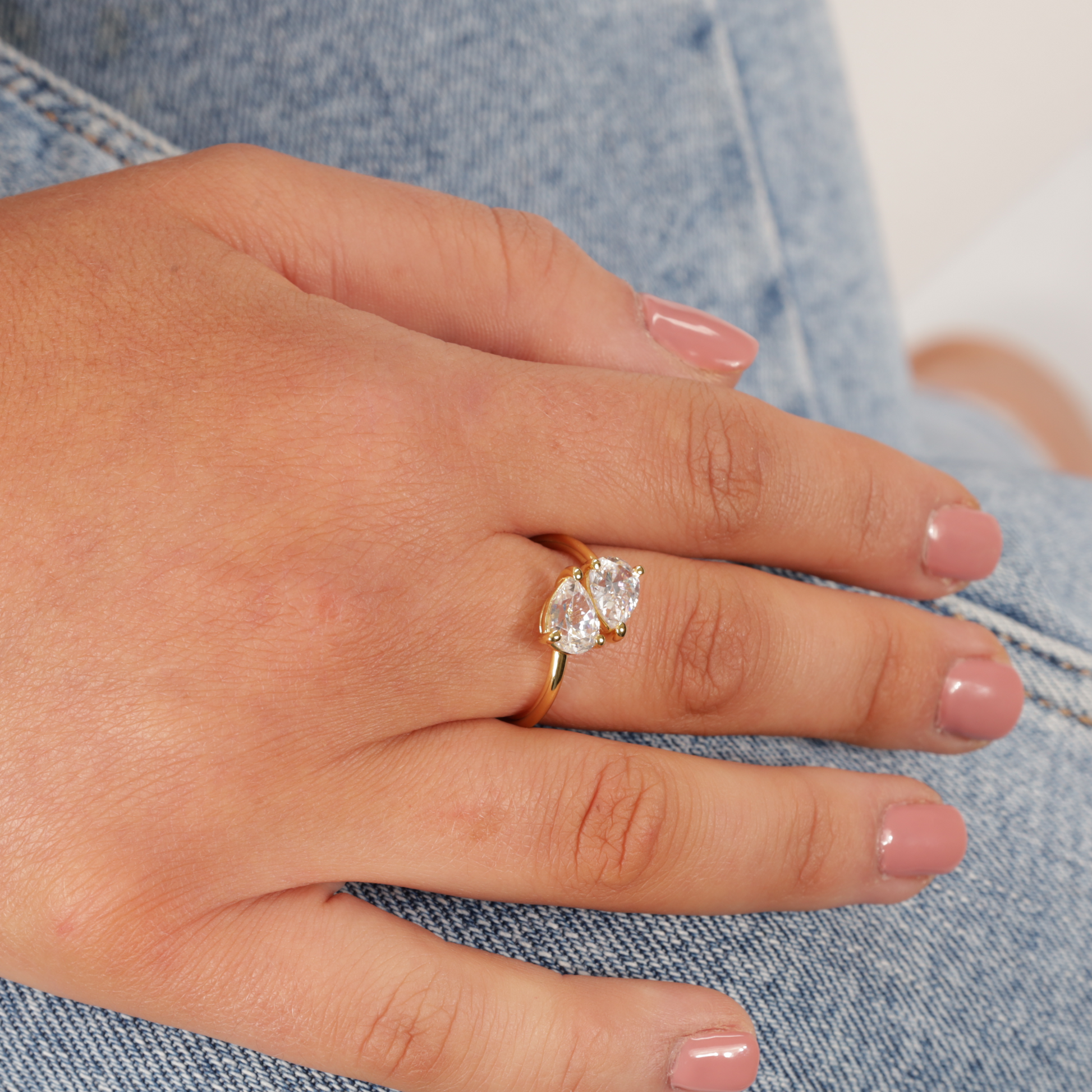 A hand with polished nails adorned with the Mia Ishaaq Gold Twist Teardrop Ring, featuring a stunning 14k gold-plated design and two large gemstones, resting on denim fabric.