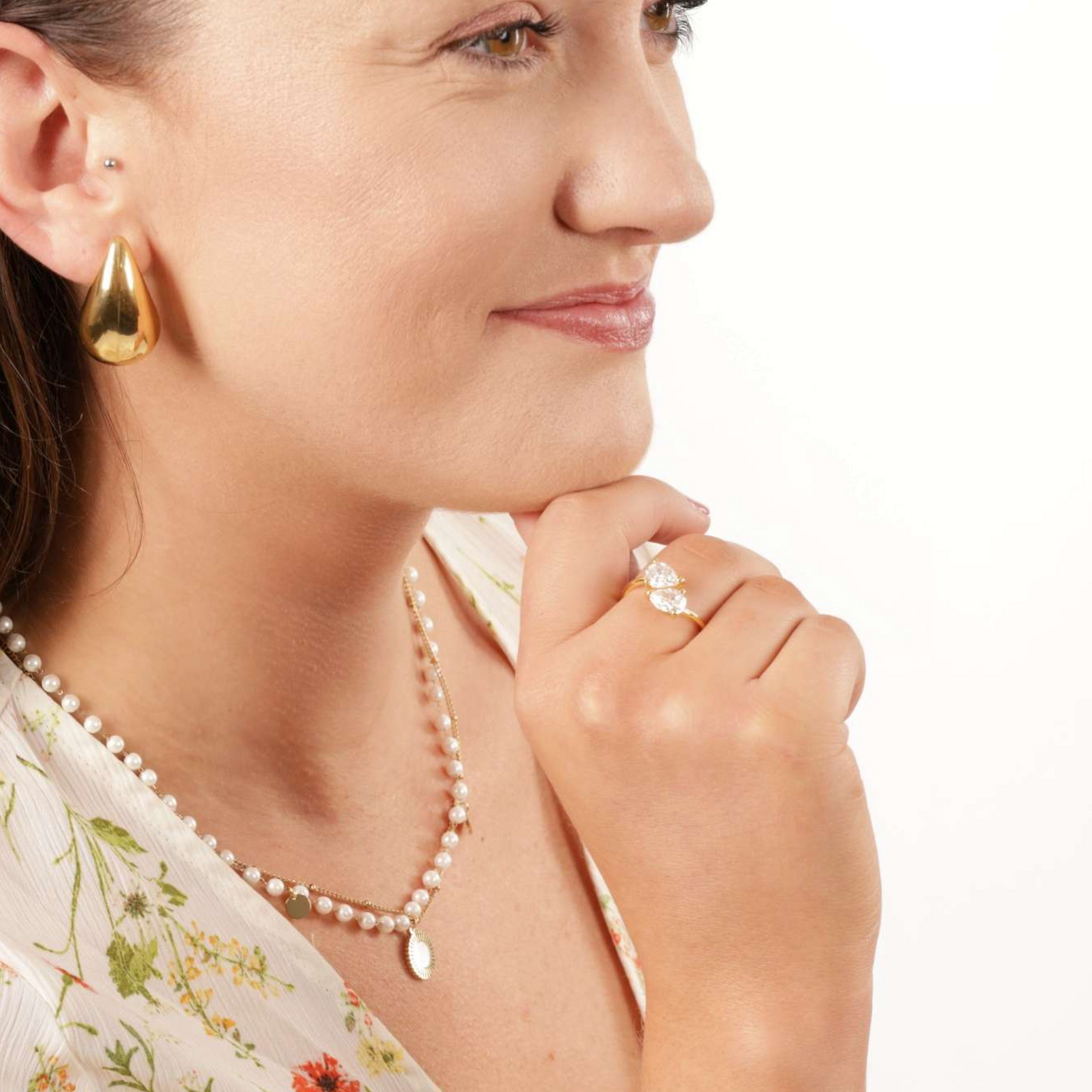 A woman with teardrop cubic zirconia earrings, a pearl necklace, and a floral top showcases the Mia Ishaaq Gold Twist Teardrop Ring on her hand against her cheek.