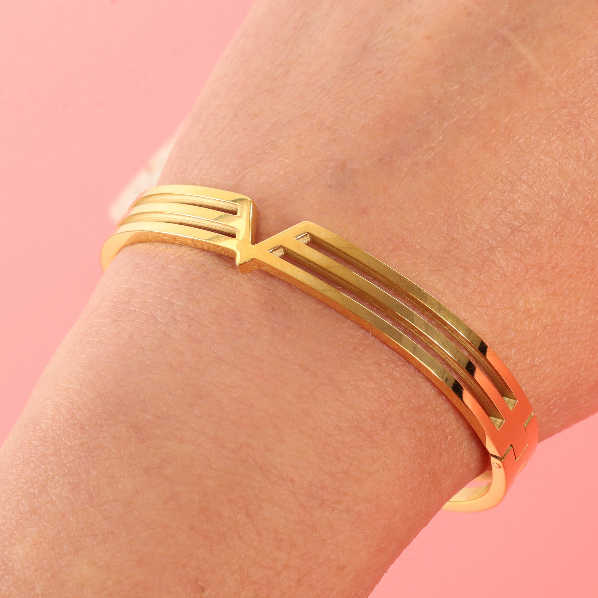 Mia Ishaaq Gold V Line Harmony Bangle Bracelet, 18k PVD Gold Plated, minimalist V-shape design, displayed on wrist against pink background, elegant jewellery for women.