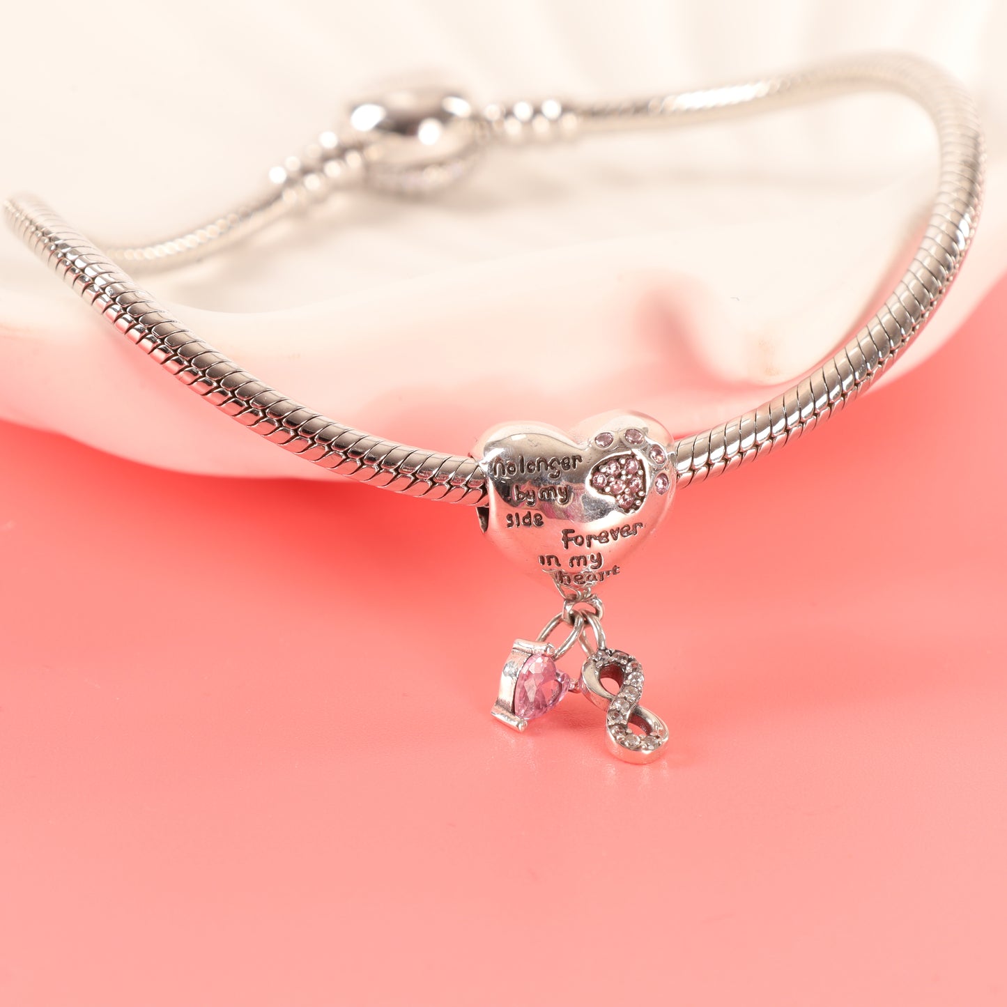 Heart-shaped dog memorial charm on a silver bracelet, draped over the edge of a white ceramic dish against a pink background - Mia Ishaaq