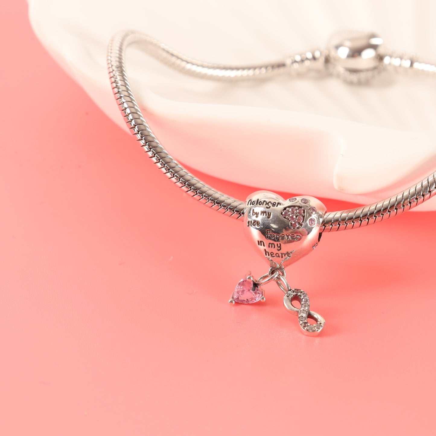 Heart-shaped dog memorial charm on a silver snake chain bracelet, hanging over the edge of a white ceramic dish against a pink background - Mia Ishaaq