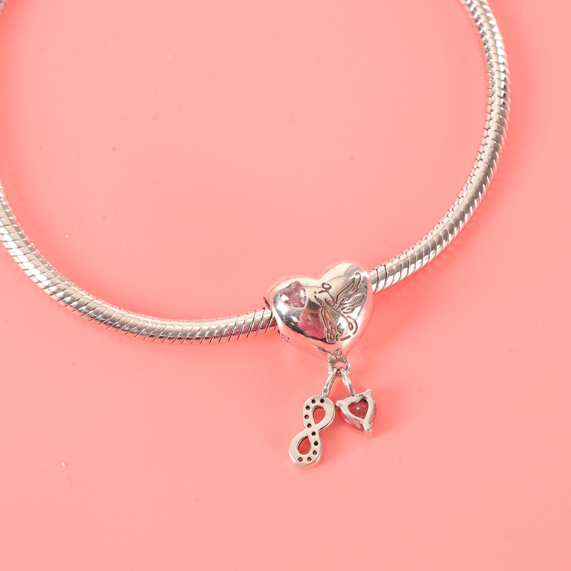 Reverse side of heart-shaped dog memorial charm with winged dog engraving and pink heart-shaped zirconia on a silver snake chain bracelet, laying on a pink background - Mia Ishaaq