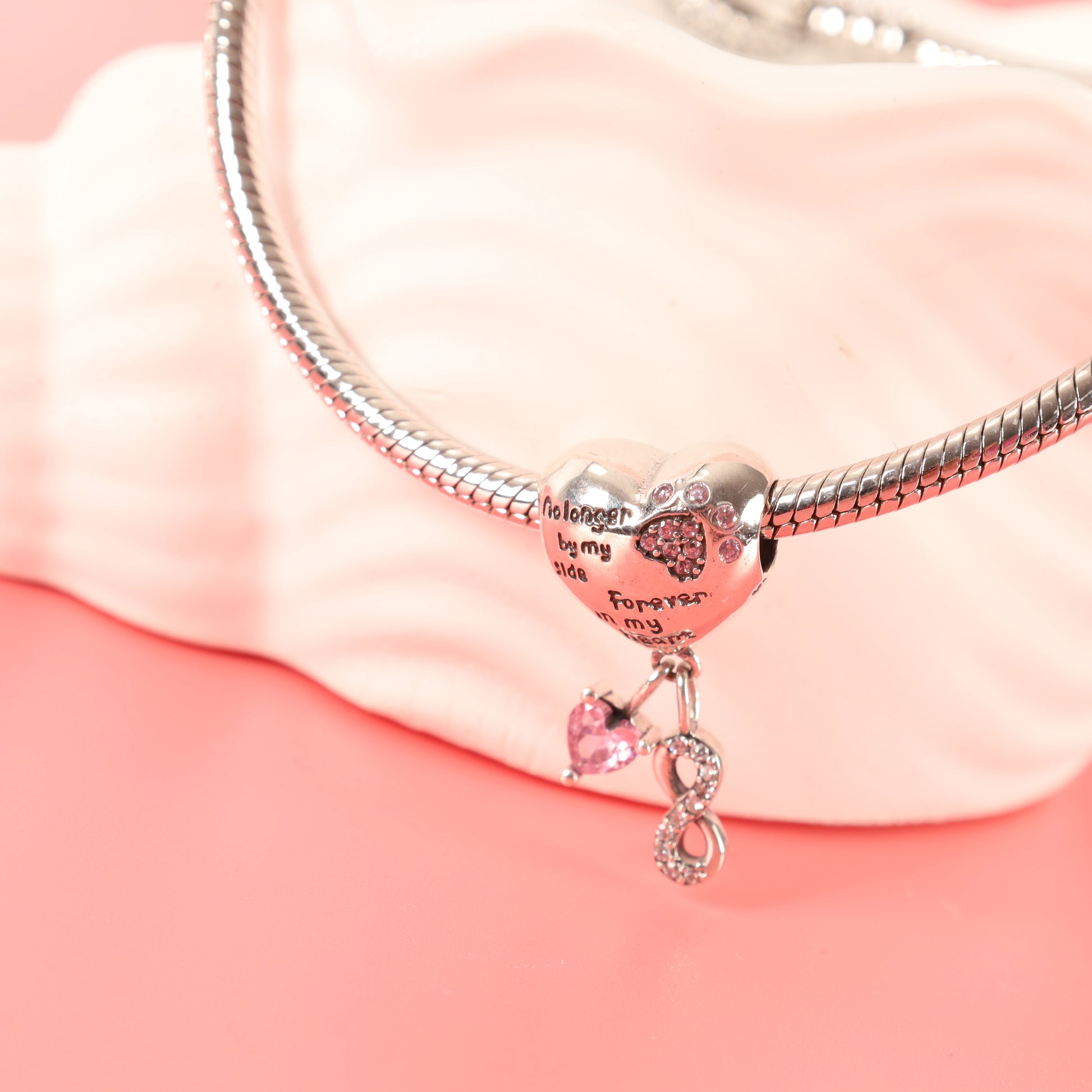 Close-up of heart-shaped dog memorial charm hanging on a silver bracelet, draped over a white ceramic dish against a pink background - Mia Ishaaq