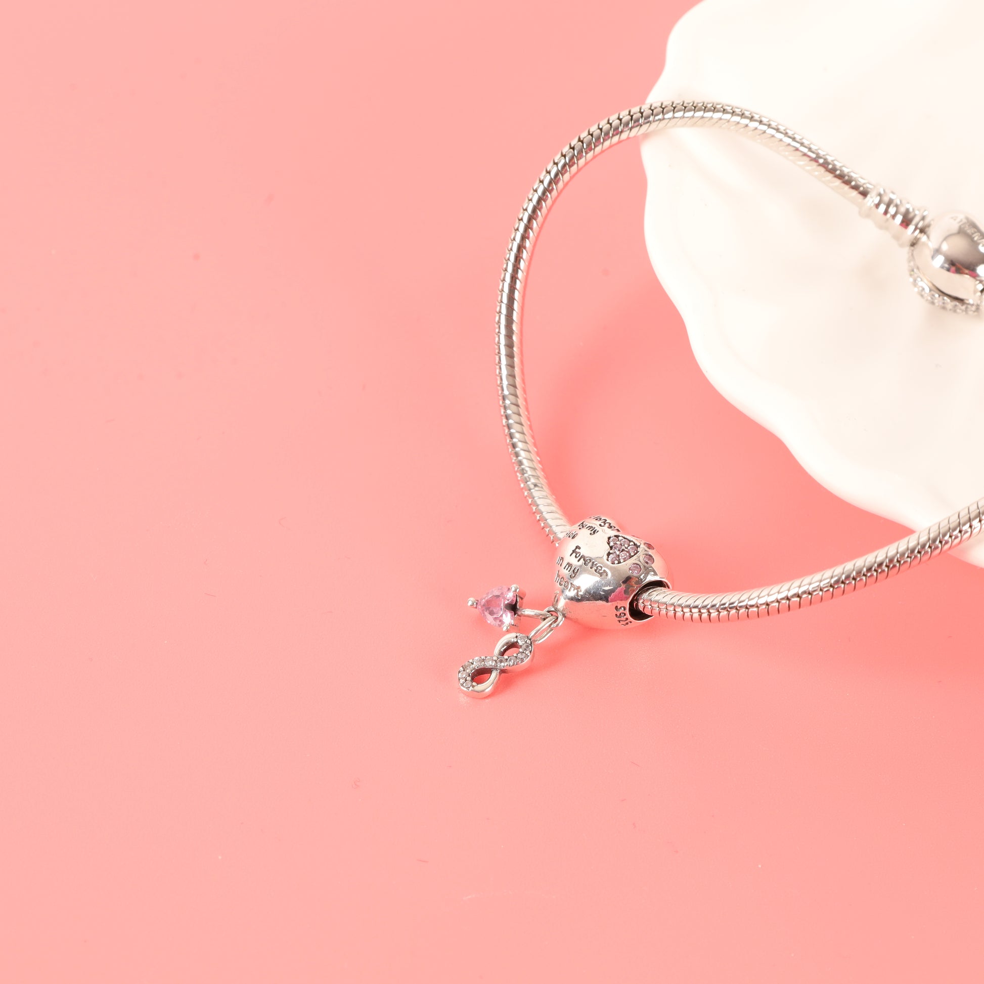 Reverse side of heart-shaped dog memorial charm with winged dog engraving and pink heart-shaped zirconia on a silver snake chain bracelet, laying next to a white ceramic dish on a pink background - Mia Ishaaq