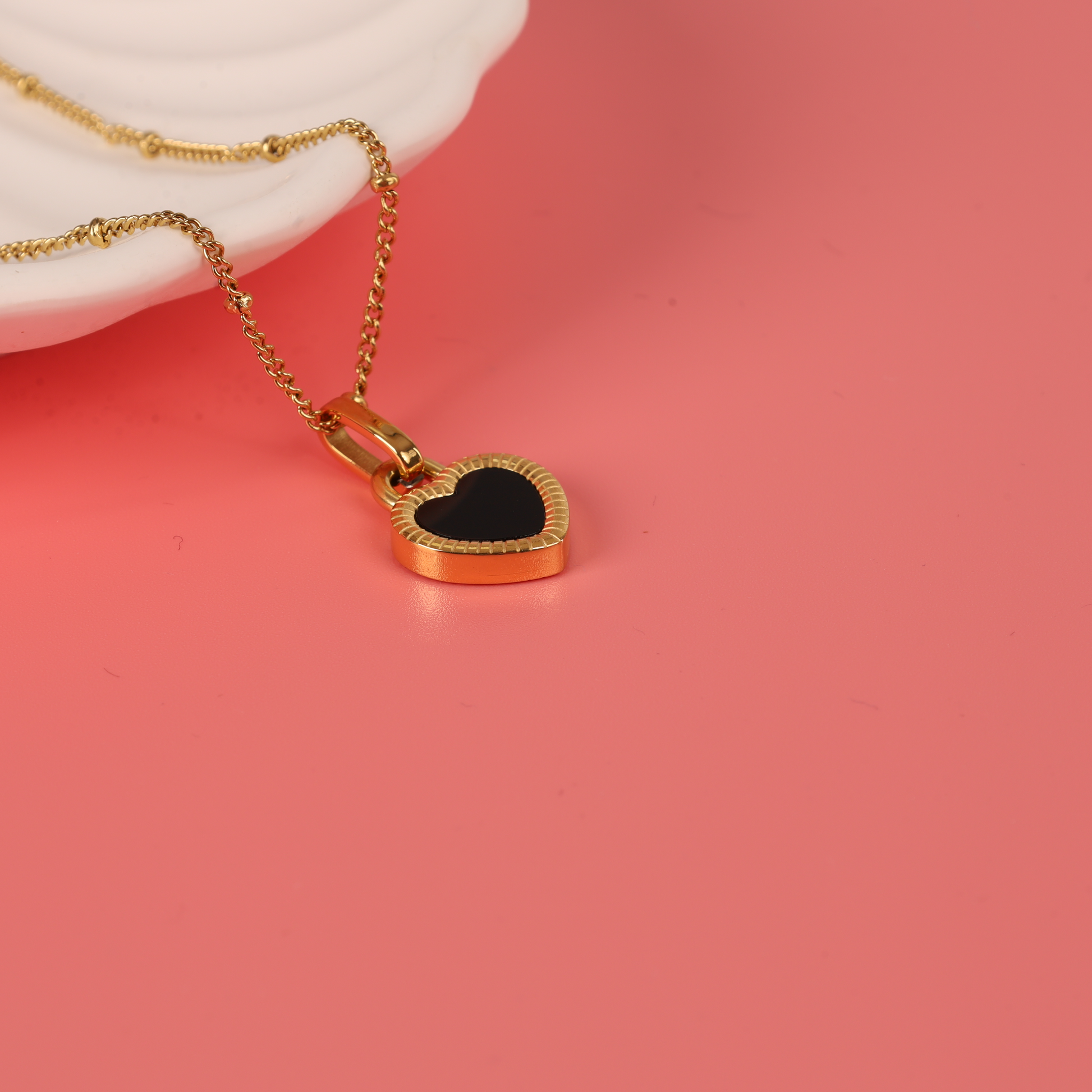 Close up side view of the Gold water-resistant reversible enamel heart pendant beaded chain necklace hanging over the edge of a white ceramic bowl on a pink background - Mia Ishaaq