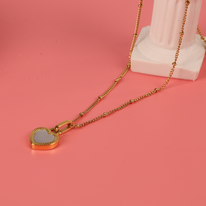 Gold water-resistant reversible enamel heart pendant beaded chain necklace placed over a white statue and laying on a pink background - Mia Ishaaq