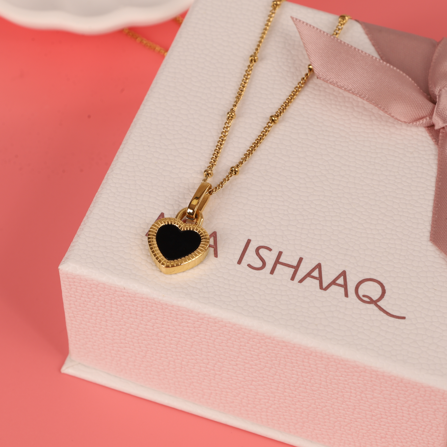 The Enamel Heart Reversible Necklace by Mia Ishaaq® elegantly rests on a pink and white gift box with a ribbon, offering versatile style perfect for any occasion.