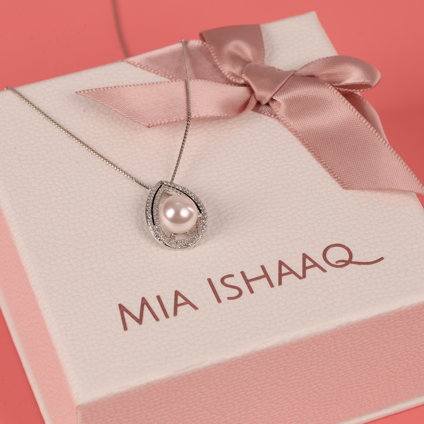 The Pearl Pendant Crystal Necklace gracefully rests on a pink gift box adorned with a delicate ribbon, featuring the elegant script of Mia Ishaaq.