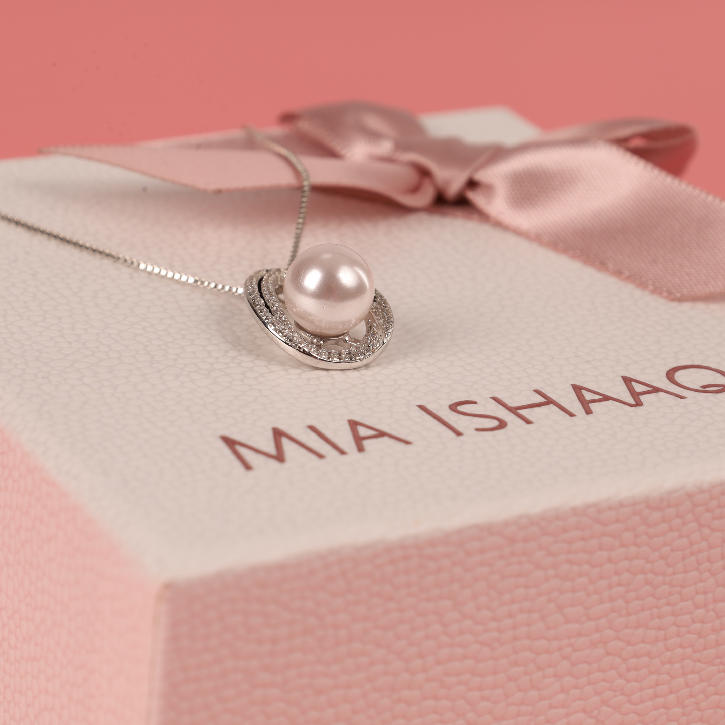 A sophisticated Pearl Pendant Crystal Necklace from Mia Ishaaq is elegantly displayed on a gift box, decorated with a pink ribbon and set against a soft pink background.