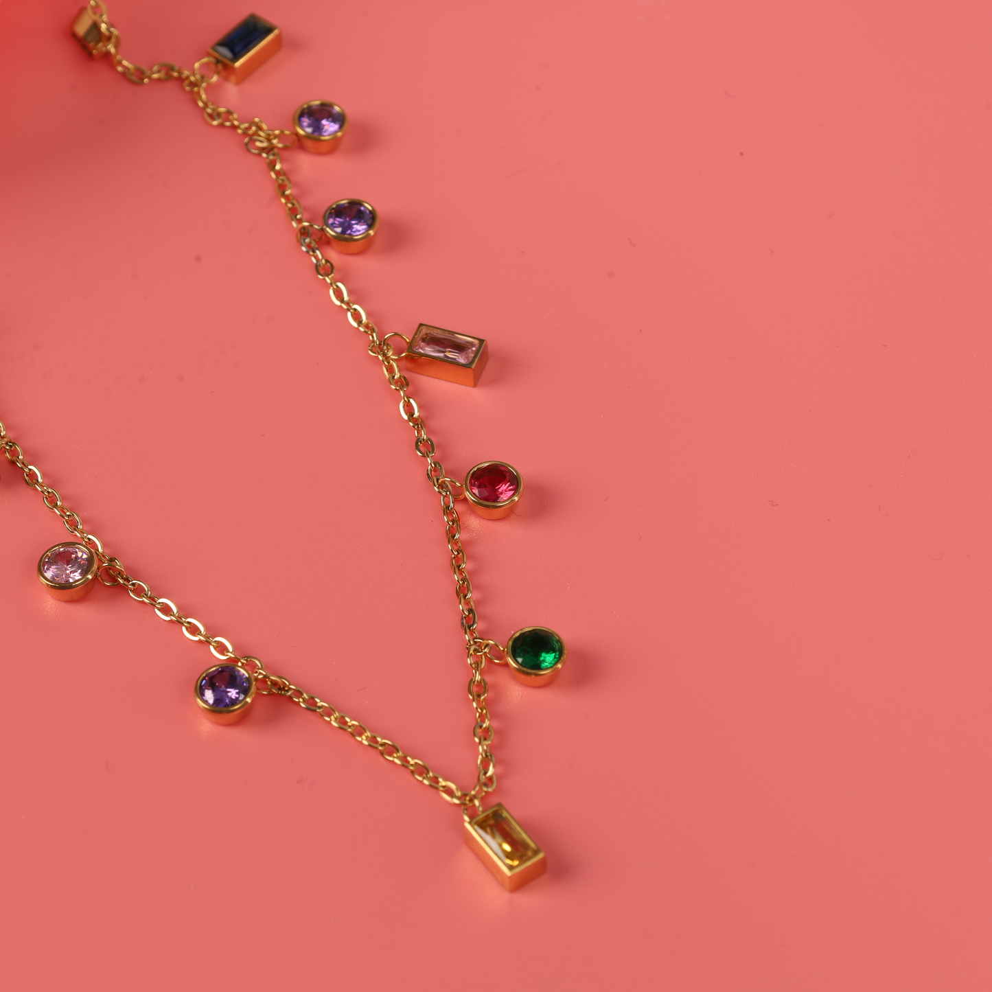 Mia Ishaaq® presents the Multicoloured Gemstone Pendant Necklace, showcasing vibrant pendants set on an 18k gold-plated chain against a coral background. Its hypoallergenic design offers both comfort and elegance.