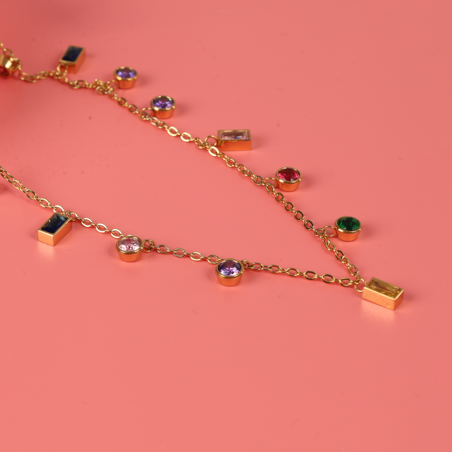 The Mia Ishaaq® Multicoloured Gemstone Pendant Necklace, plated in 18k gold and featuring vibrant gemstone pendants, is displayed against a pink background. This piece is an ideal hypoallergenic option for jewelry enthusiasts.