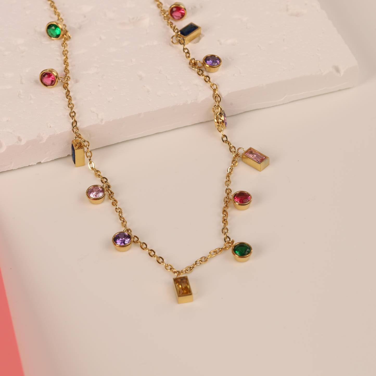 The Multicoloured Gemstone Pendant Necklace by Mia Ishaaq® boasts a stunning array of vibrant stones elegantly arranged on a white and pink surface. Crafted with luxurious 18k gold plated metal, its hypoallergenic design ensures maximum comfort for even the most sensitive skin.