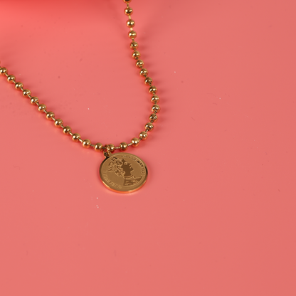 The Beaded Chain Coin Pendant Necklace by Mia Ishaaq showcases a breathtaking 18k gold-plated coin on a beaded chain, exquisitely displayed against a coral pink background.