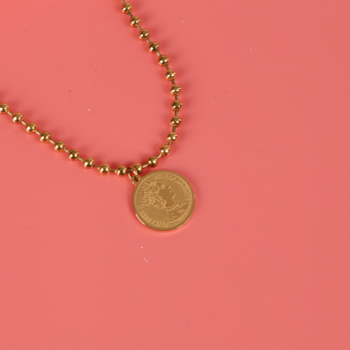 The Beaded Chain Coin Pendant Necklace by Mia Ishaaq, featuring an 18k gold-plated coin, elegantly hangs against a delicate pink background.