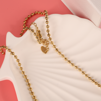 The Beaded Chain Coin Pendant Necklace by Mia Ishaaq, featuring an 18k gold-plated beaded design and a heart charm, gracefully rests on a white shell-shaped dish against a pink background.