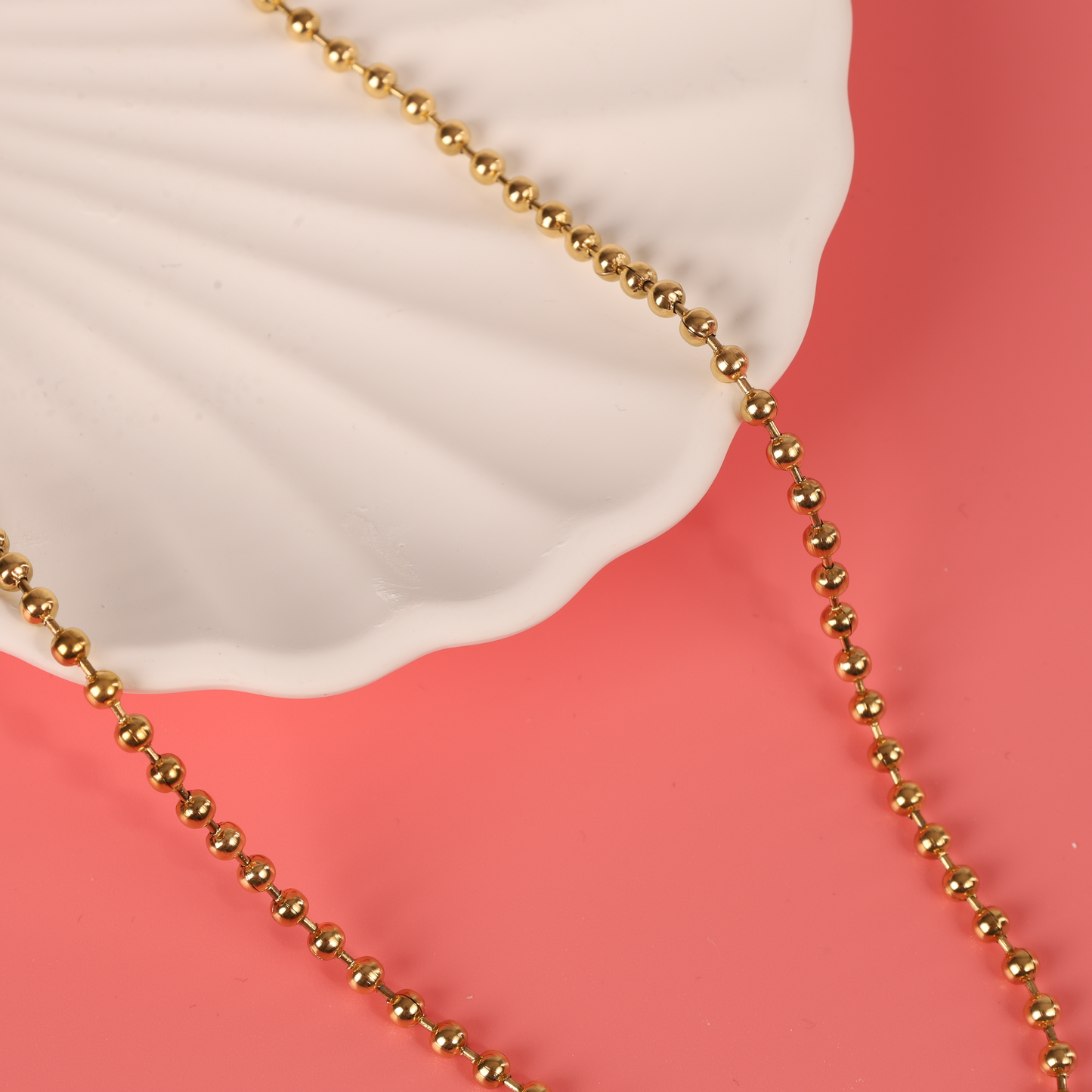 A Beaded Chain Coin Pendant Necklace by Mia Ishaaq, featuring 18k gold plating, gracefully rests over a white shell-shaped dish set against a pink backdrop.