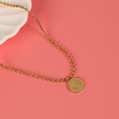 The Beaded Chain Coin Pendant Necklace by Mia Ishaaq, featuring an elegant 18k gold-plated chain with a round pendant, is beautifully displayed over a white shell on a soft pink background.