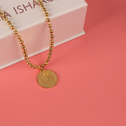 The Beaded Chain Coin Pendant Necklace by Mia Ishaaq, featuring a vibrant coin pendant with 18k gold plating, is elegantly displayed on a soft pink surface next to a pristine white box.