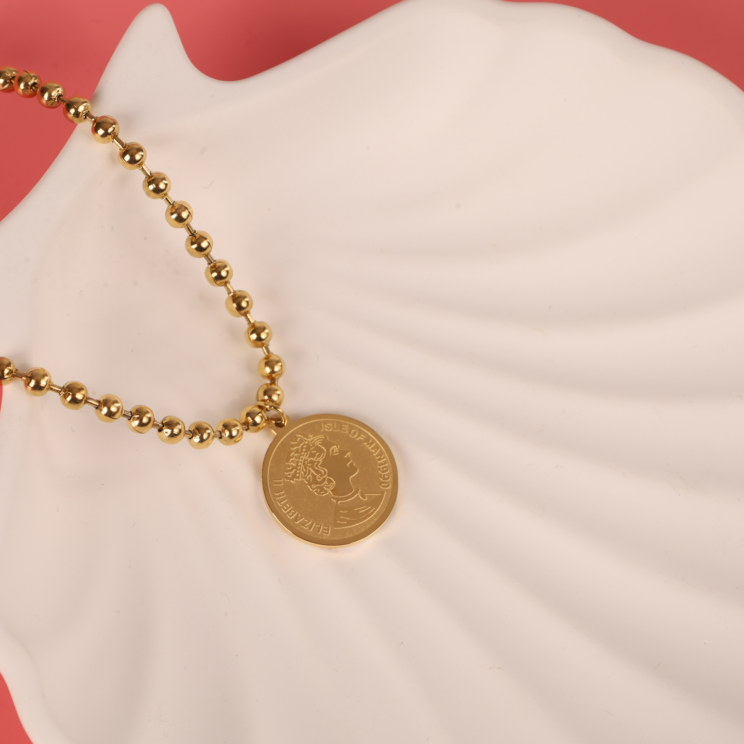 A beautifully crafted Beaded Chain Coin Pendant Necklace by Mia Ishaaq, featuring 18k gold plating, is gracefully placed on a white shell-shaped dish against a soft pink background.