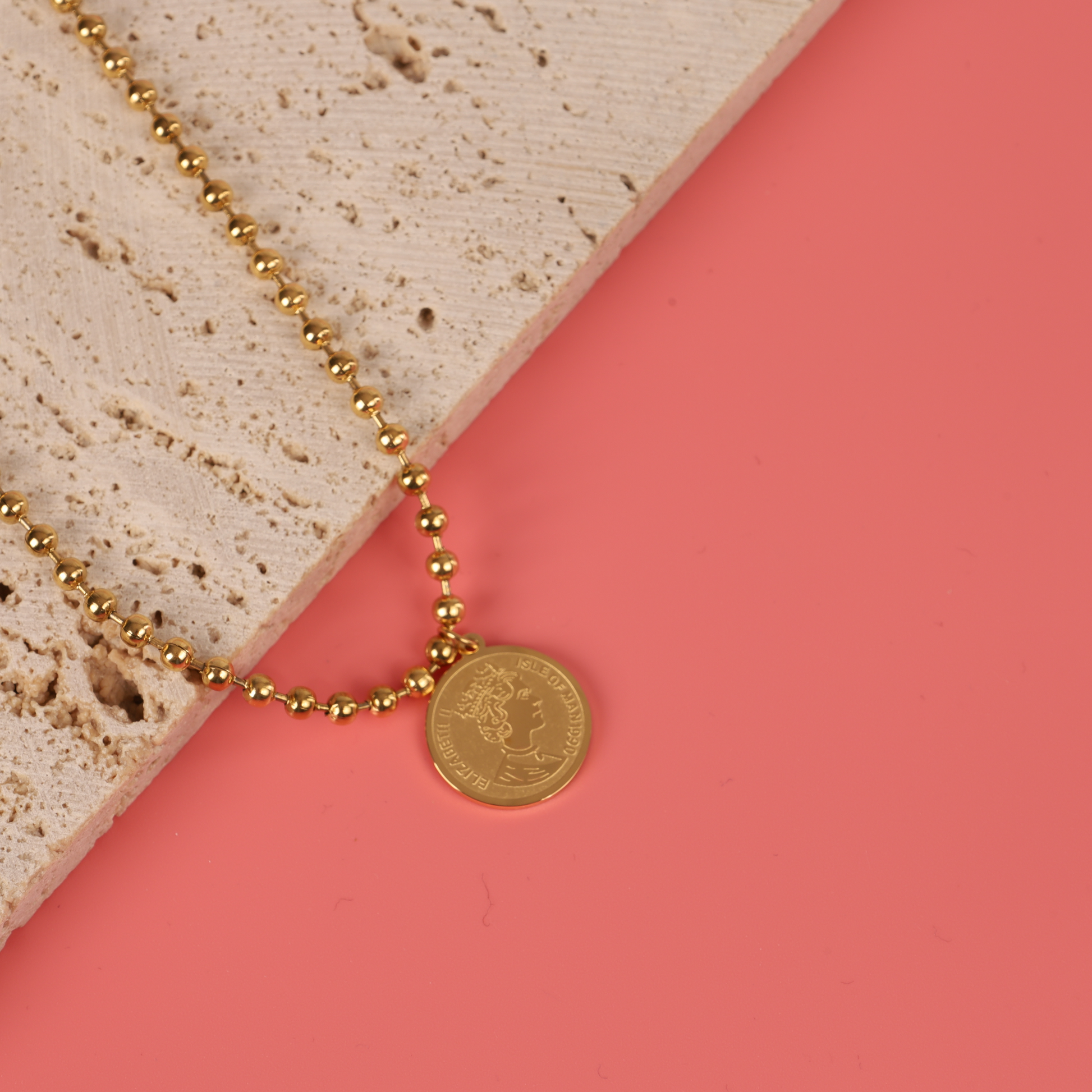 Mia Ishaaq's Beaded Chain Coin Pendant Necklace, 18k gold plated, displayed on a stone slab set against a coral background.