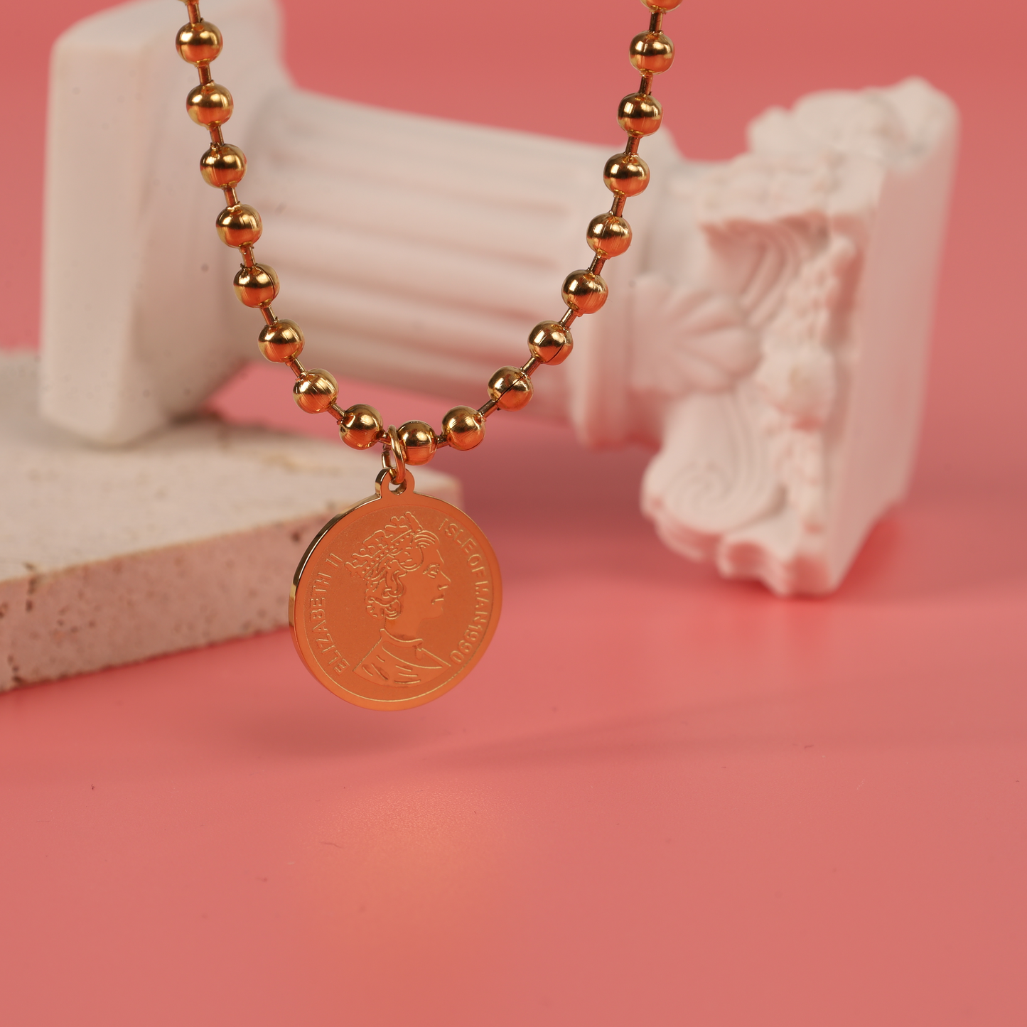 The Beaded Chain Coin Pendant Necklace by Mia Ishaaq shines with its 18k gold plating, set beautifully on a pink backdrop and encircled by white classical columns.