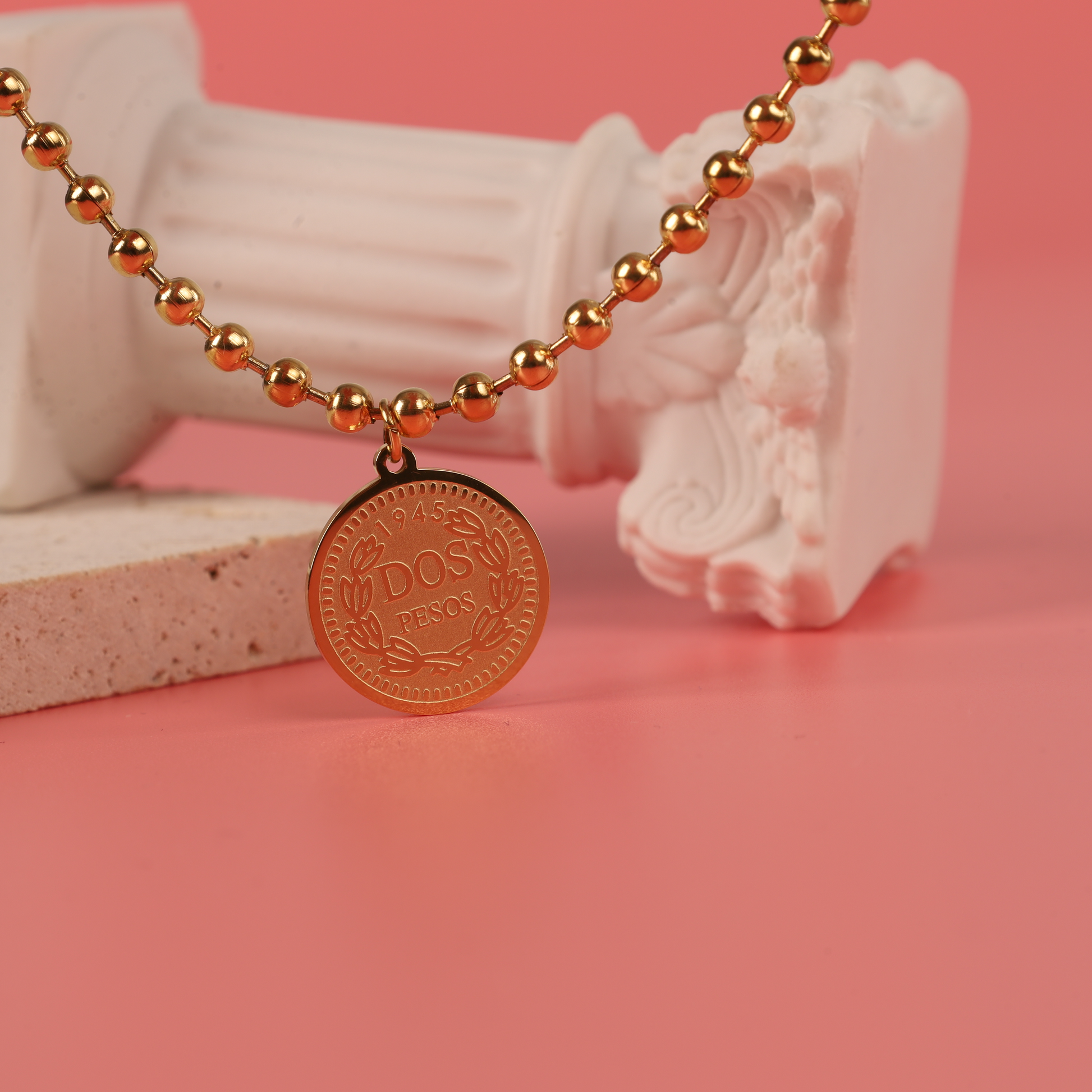 The Beaded Chain Coin Pendant Necklace by Mia Ishaaq is elegantly showcased on decorative marble pieces against a pink backdrop, featuring luxurious 18k gold plating and a stunning Dos Pesos coin pendant.