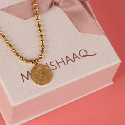 The Beaded Chain Coin Pendant Necklace by Mia Ishaaq, with its 18k gold plating, captivates the eye as it rests on a textured white box adorned with a pink ribbon, all beautifully set against a soft pink background.